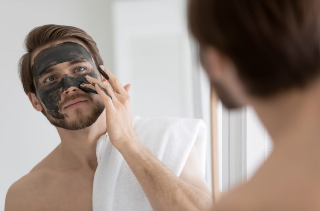 Young man applying black charcoal clay mask on face at home